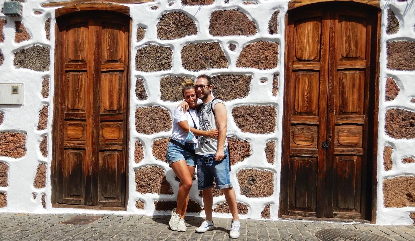 Pareja abrazada ante un muro de piedra volcánica y puertas de madera en un pueblo de Gran Canaria