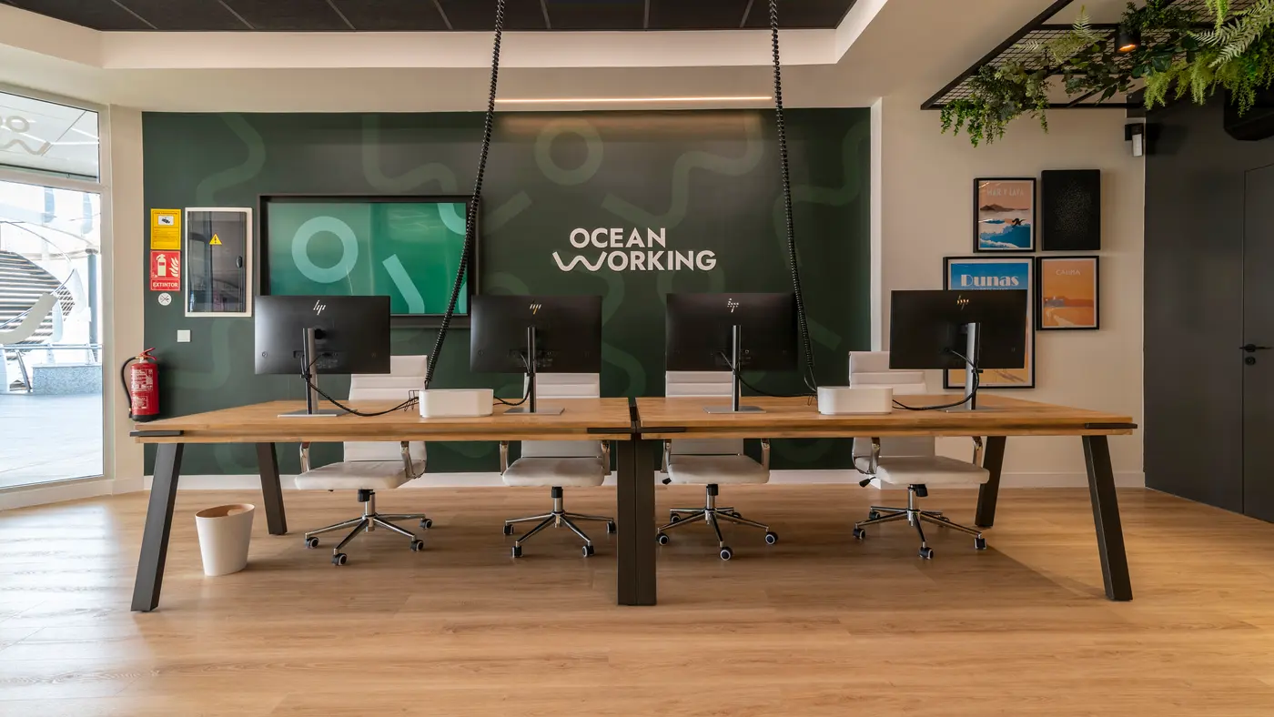 Mesa de coworking con cuatro monitores frente a la pared verde con el logo de OceanWorking