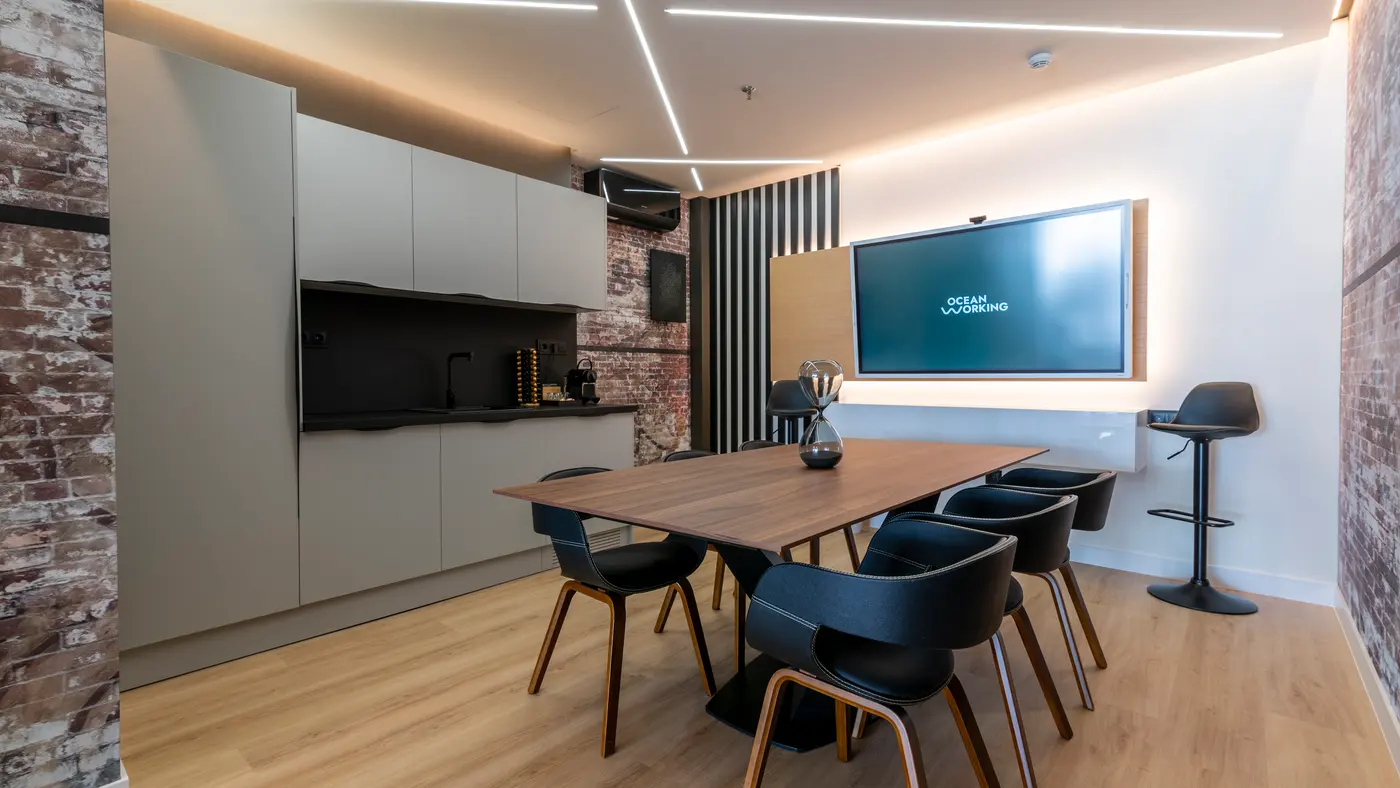 Sala de reuniones con mesa de nogal, sillas negras y gran pantalla, junto a un office con pared de ladrillo