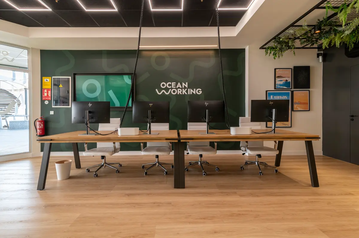 Sala de coworking de OceanWorking con escritorios de madera, monitores y la pared verde con el logo