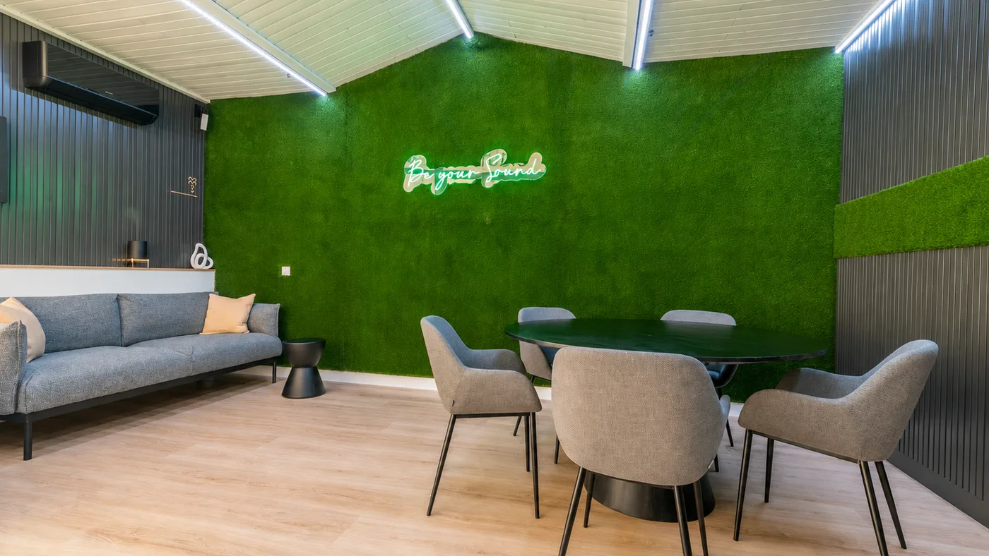 Estudio privado con sofá gris, mesa con sillas y pared de césped verde con rótulo de neón