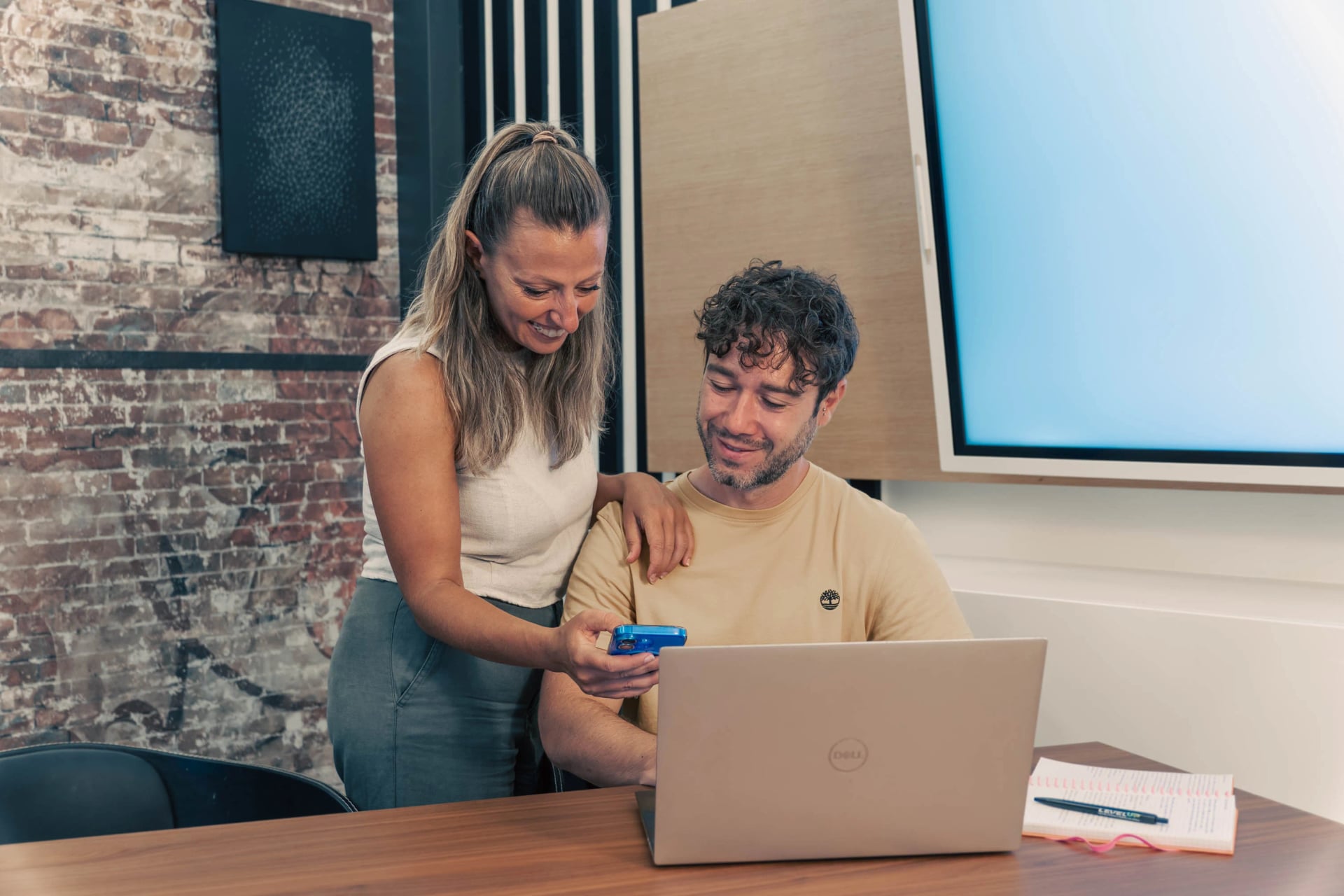 Erika y Matteo en el coworking de Oceanworking en Maspalomas, Gran Canaria: ella, de pie, le muestra algo en el móvil mientras él trabaja con el portátil, frente a la pared de ladrillo visto.