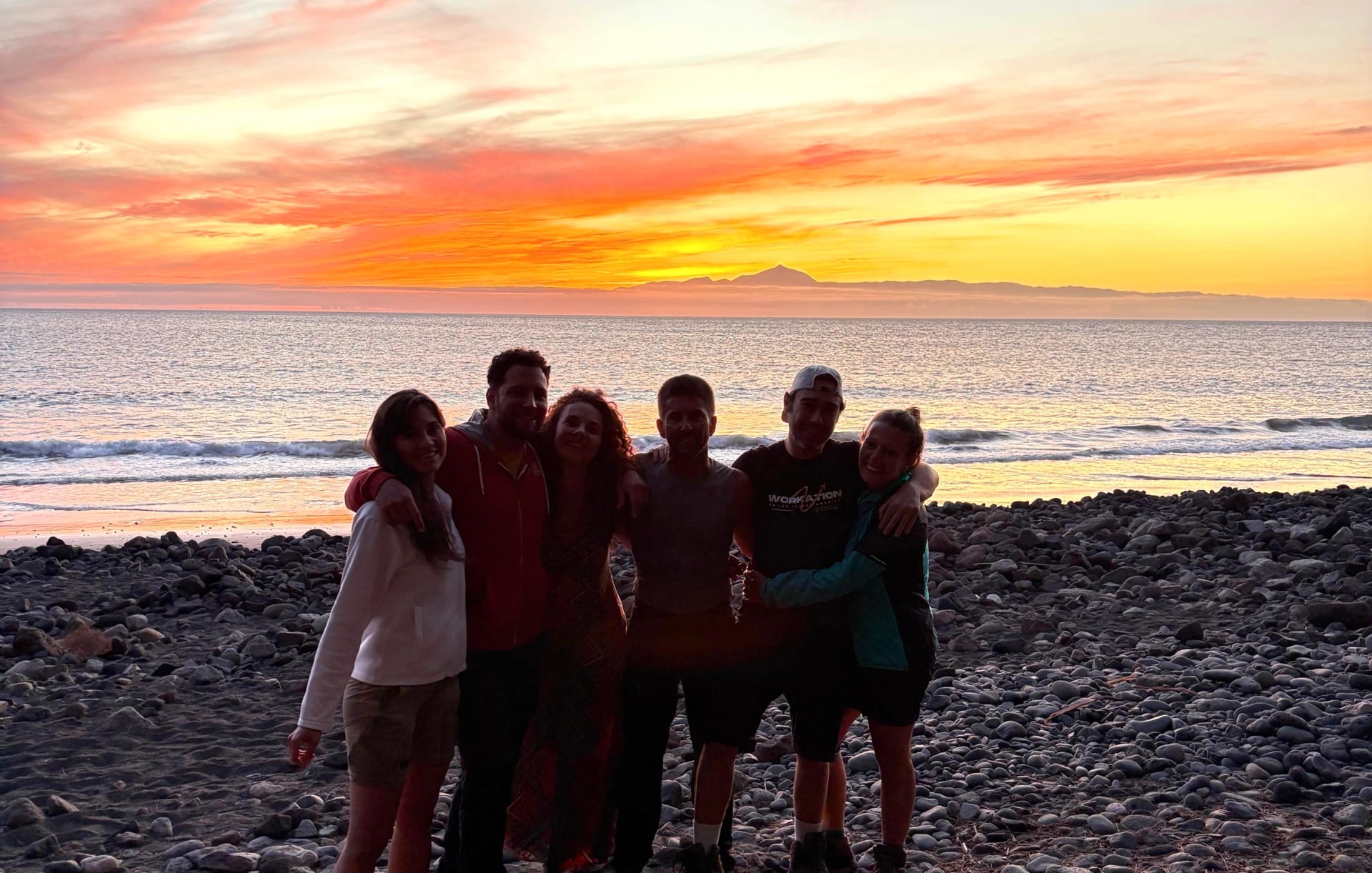 Grupo de amigos abrazados en una playa de cantos rodados al atardecer, con el océano y la silueta del Teide al fondo, en Gran Canaria
